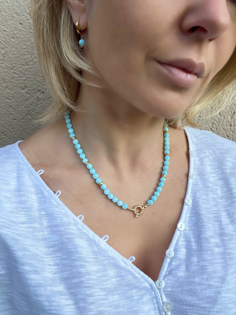 Op de afbeelding: Een lichtblauwe kralenketting met een gouden sluiting. De ketting is gemaakt van kleine, ronde kralen en wordt gedragen door een persoon met blond haar.