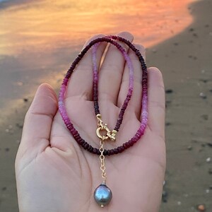 May include: A delicate necklace made with pink and red gemstone beads, featuring a gold clasp and a single black pearl pendant.
