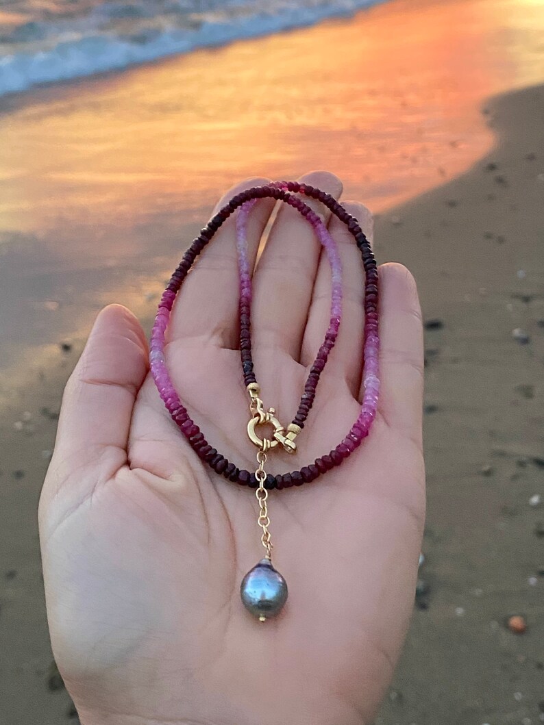 May include: A delicate necklace made with pink and red gemstone beads, featuring a gold clasp and a single black pearl pendant.
