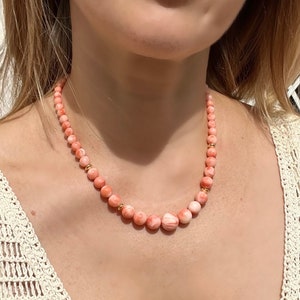 May include: A delicate necklace made of pink coral beads with gold accents. The necklace is worn by a person with a white crocheted top.