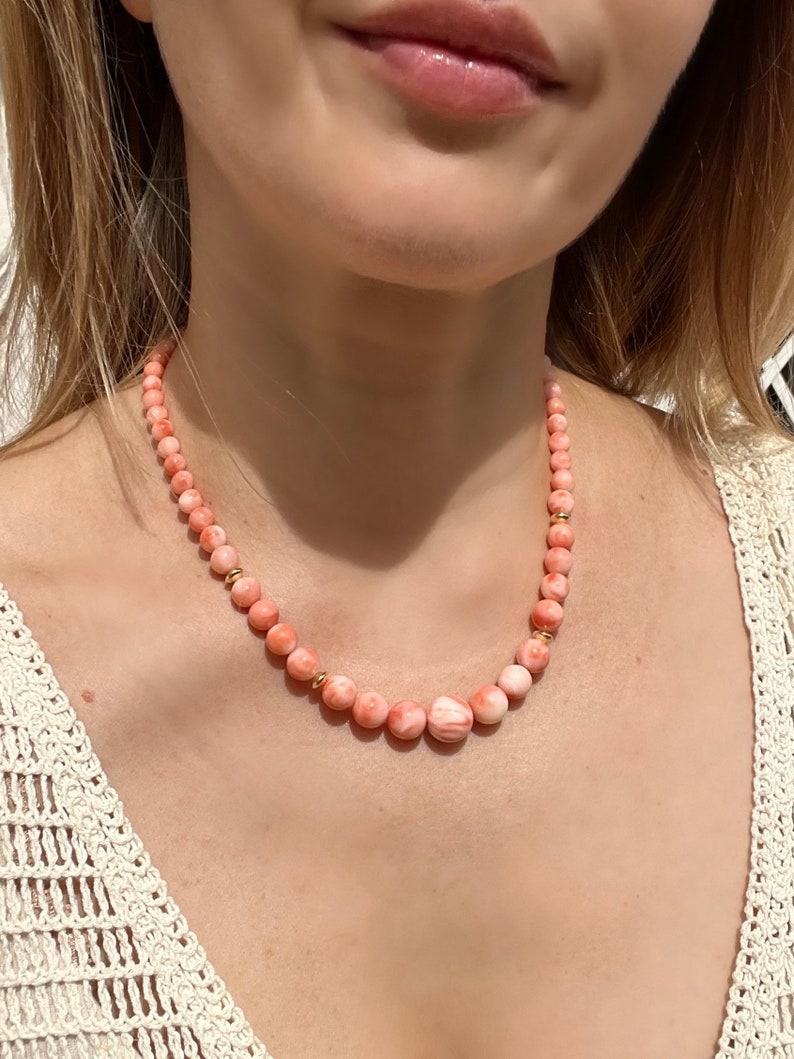 May include: A delicate necklace made of pink coral beads with gold accents. The necklace is worn by a person with a white crocheted top.