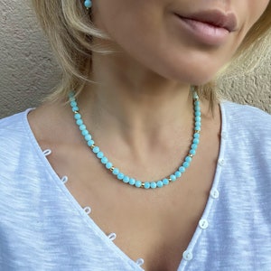 Op de afbeelding: Een lichtblauwe kralenketting met gouden accenten. De ketting is gemaakt van kleine, ronde kralen en wordt gedragen door een persoon met blond haar.