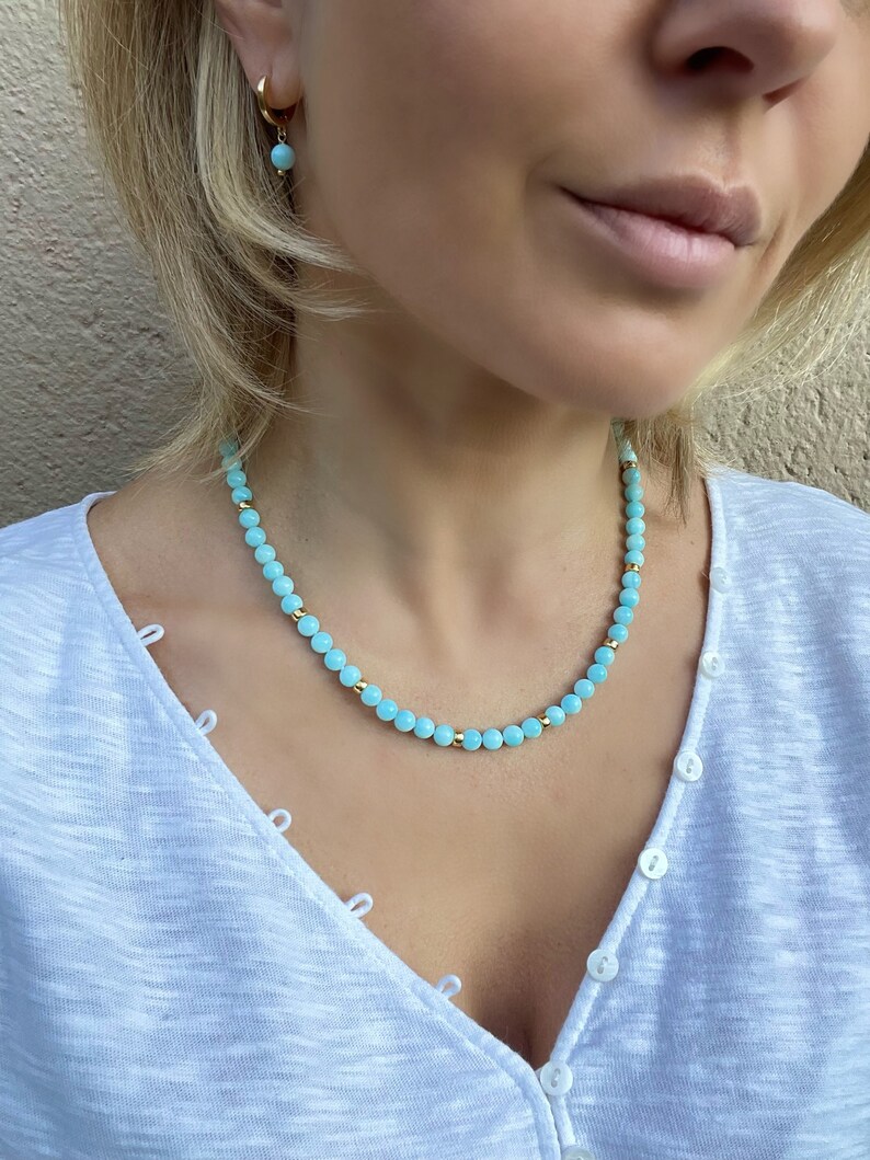 Op de afbeelding: Een lichtblauwe kralenketting met gouden accenten. De ketting is gemaakt van kleine, ronde kralen en wordt gedragen door een persoon met blond haar.