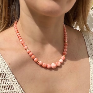 May include: A delicate necklace made of pink and white coral beads with gold accents. The necklace is worn by a person with a white crocheted top.