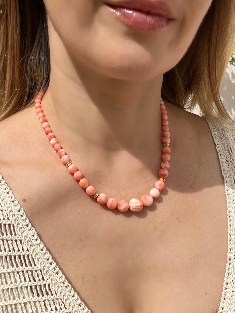 May include: A delicate necklace made of pink and white coral beads with gold accents. The necklace is worn by a person with a white crocheted top.