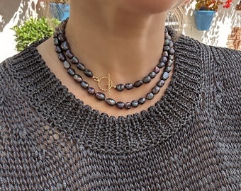 Può includere: Una donna indossa un maglione grigio scuro con una collana a doppio filo di perle nere con una chiusura dorata.