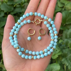 Op de afbeelding: Een lichtblauwe edelsteen ketting en bijpassende oorbellen. De ketting is gemaakt van kleine, ronde kralen met gouden accenten. De oorbellen zijn hoepelvormig met een enkele kraal die aan elke hoepel hangt.