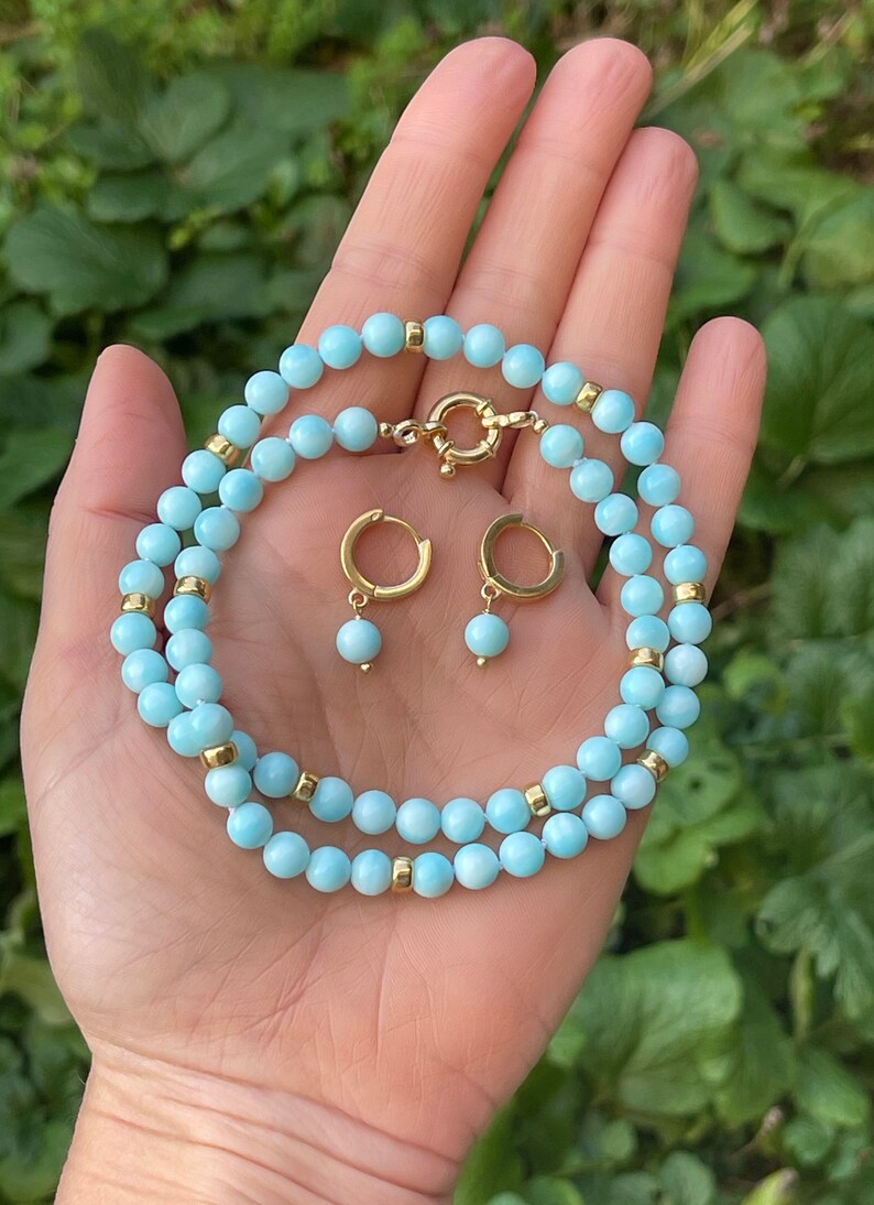 Op de afbeelding: Een lichtblauwe edelsteen ketting en bijpassende oorbellen. De ketting is gemaakt van kleine, ronde kralen met gouden accenten. De oorbellen zijn hoepelvormig met een enkele kraal die aan elke hoepel hangt.