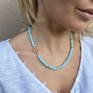 Op de afbeelding: Een lichtblauwe kralenketting met een gouden sluiting. De ketting is gemaakt van kleine, ronde kralen en wordt gedragen door een persoon met een wit shirt.