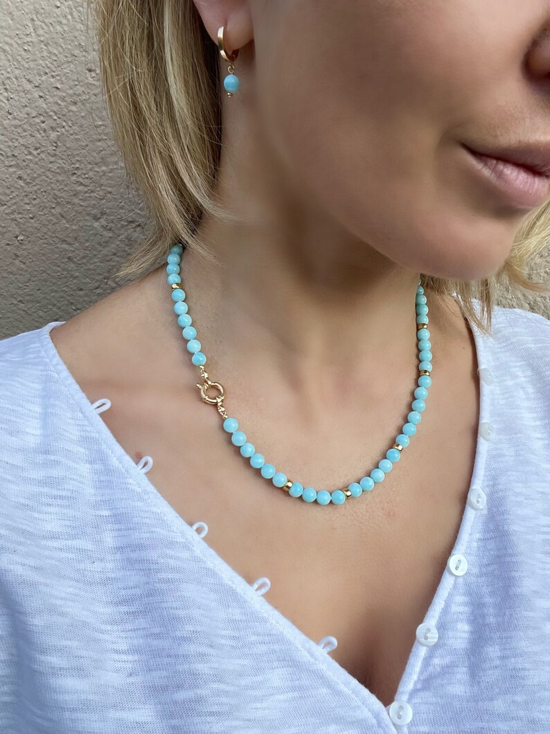 Op de afbeelding: Een lichtblauwe kralenketting met een gouden sluiting. De ketting is gemaakt van kleine, ronde kralen en wordt gedragen door een persoon met een wit shirt.