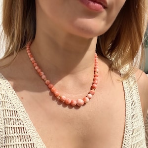 May include: A pink and white coral bead necklace with gold accents. The necklace is worn by a person with a white crocheted top.