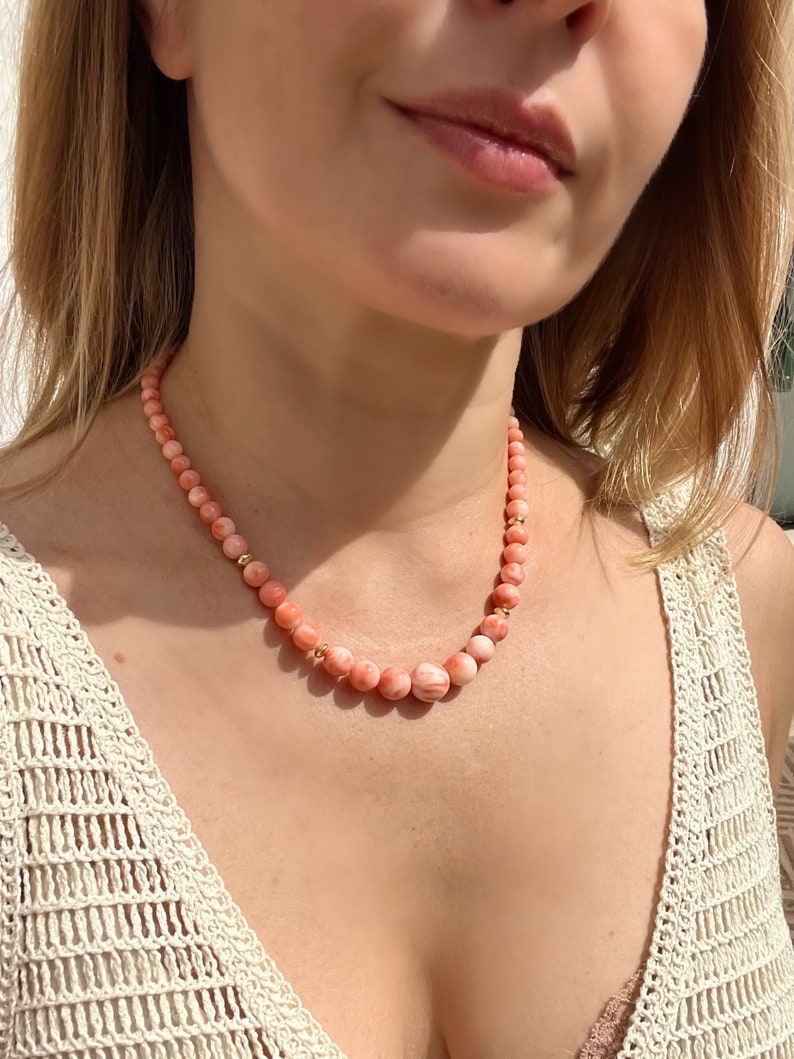 May include: A pink and white coral bead necklace with gold accents. The necklace is worn by a person with a white crocheted top.