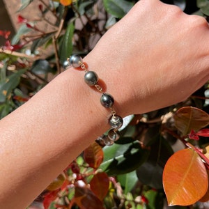 May include: A gold chain bracelet with dark grey pearls. The bracelet is on a person's wrist.