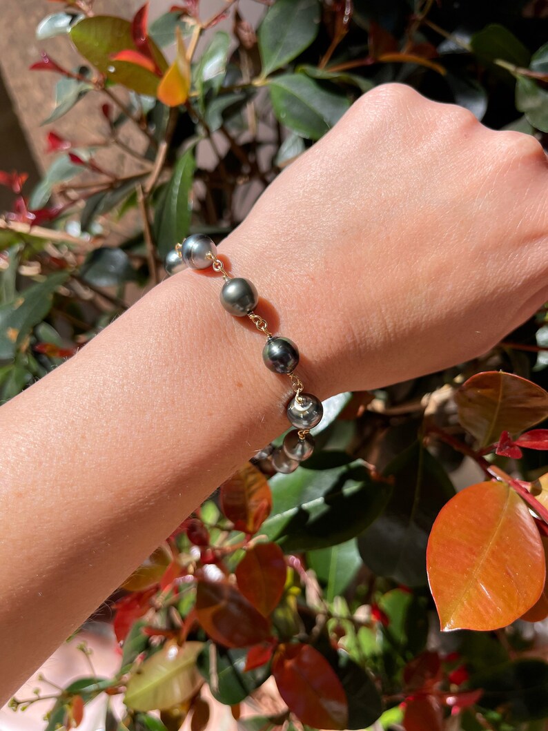May include: A gold chain bracelet with dark grey pearls. The bracelet is on a person's wrist.