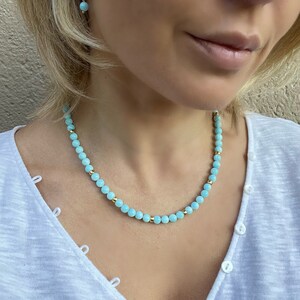 Op de afbeelding: Een lichtblauwe kralenketting met gouden accenten. De ketting is gemaakt van kleine, ronde kralen en wordt gedragen door een persoon met blond haar.