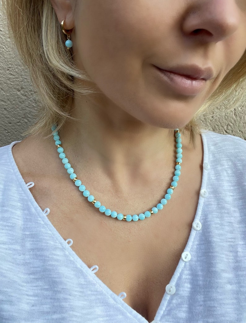Op de afbeelding: Een lichtblauwe kralenketting met gouden accenten. De ketting is gemaakt van kleine, ronde kralen en wordt gedragen door een persoon met blond haar.