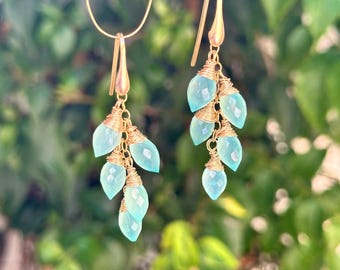 Puede incluir: Un par de pendientes de oro con gotas de piedras preciosas de color azul claro. Los pendientes están hechos con alambre envuelto alrededor de las piedras preciosas, creando un efecto en cascada.