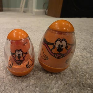 May include: Two vintage orange and clear plastic toys featuring a cartoon dog character. The toys have an egg-like shape with a rounded top and base. The dog character is printed on the front of the toys.