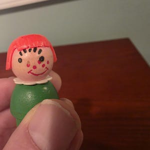 May include: A vintage wooden toy figure with a painted face, red hair, and a green body. The figure has a smiling face with black eyes and red cheeks. The toy is held in a hand.