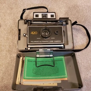 May include: A black Polaroid Land Camera model 420 with a green instruction booklet inside. The camera has a black strap and a silver lens.