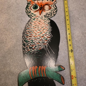 May include: A flat, cutout-style owl wall decoration featuring black, orange, and green colors. The owl is perched on a branch and measures approximately 18 inches in height when compared to the yellow measuring tape placed alongside it.