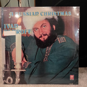 May include: A vinyl record cover titled A RUSSIAN CHRISTMAS by IVAN REBROFF. The cover features a bearded individual wearing a white fur hat and a dark green tunic with decorative trim, positioned next to a tall white candle in a silver holder.