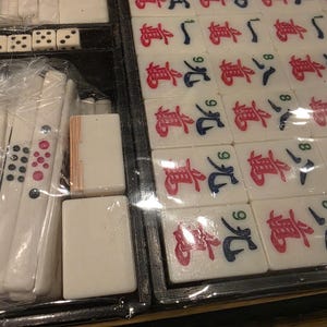 Może przedstawiać: Zbliżenie zestawu do gry w Mahjong. Zestaw zawiera białe kafelki z czerwonymi i niebieskimi symbolami, kości do gry i inne elementy gry. Kafelki są ułożone w czarnej tacy, a niekt&oacute;re elementy są owinięte w plastik.