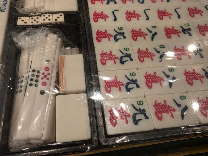 Może przedstawiać: Zbliżenie zestawu do gry w Mahjong. Zestaw zawiera białe kafelki z czerwonymi i niebieskimi symbolami, kości do gry i inne elementy gry. Kafelki są ułożone w czarnej tacy, a niekt&oacute;re elementy są owinięte w plastik.