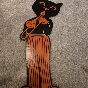 May include: A flat, die-cut cardboard decoration featuring a black cat playing a trombone. The cat has orange eyes, nose, and striped pants. The base includes the text "MADE IN U.S.A." and "Copr. H. E. Luhrs" printed in small black lettering.