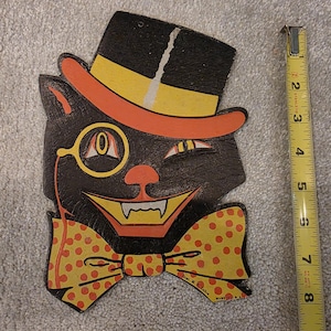 May include: A black cat cutout decoration wearing a black top hat with an orange band, a yellow monocle, and a yellow bowtie with orange polka dots. The cat has a wide grin with visible white fangs. A yellow measuring tape shows a height of 8 inches.