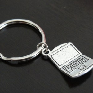 May include: Silver-colored laptop charm keychain. The charm is a detailed miniature of a laptop computer, complete with a keyboard and screen. The keychain has a silver split ring.