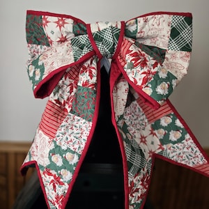 May include: A large, quilted Christmas bow with a patchwork design. The bow features red, green, and white fabrics with festive patterns, including poinsettias, stars, and Santa Claus. The edges are trimmed with red binding.