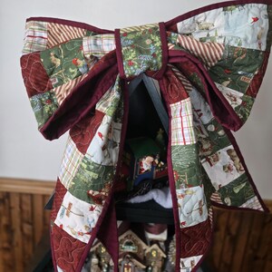 May include: A large, quilted Christmas bow in shades of red, green, and white, with a dark red border. The bow is made of patchwork fabric and is draped over a black shelf holding a miniature Christmas village and a toy train.