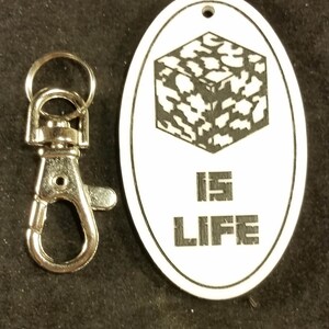 以下が含まれることがあります： 白地に黒と白の迷彩柄の立方体と「IS LIFE」の文字が書かれた白い楕円形のキーホルダーです。