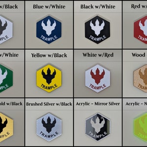 May include: A set of 12 hexagonal game tokens with a dinosaur footprint design. The tokens are made in various materials and colors, including white, black, blue, red, green, yellow, wood, brushed gold, brushed silver, mirror silver, and neon green. Each token has the word "TRAMPLE" printed on it.