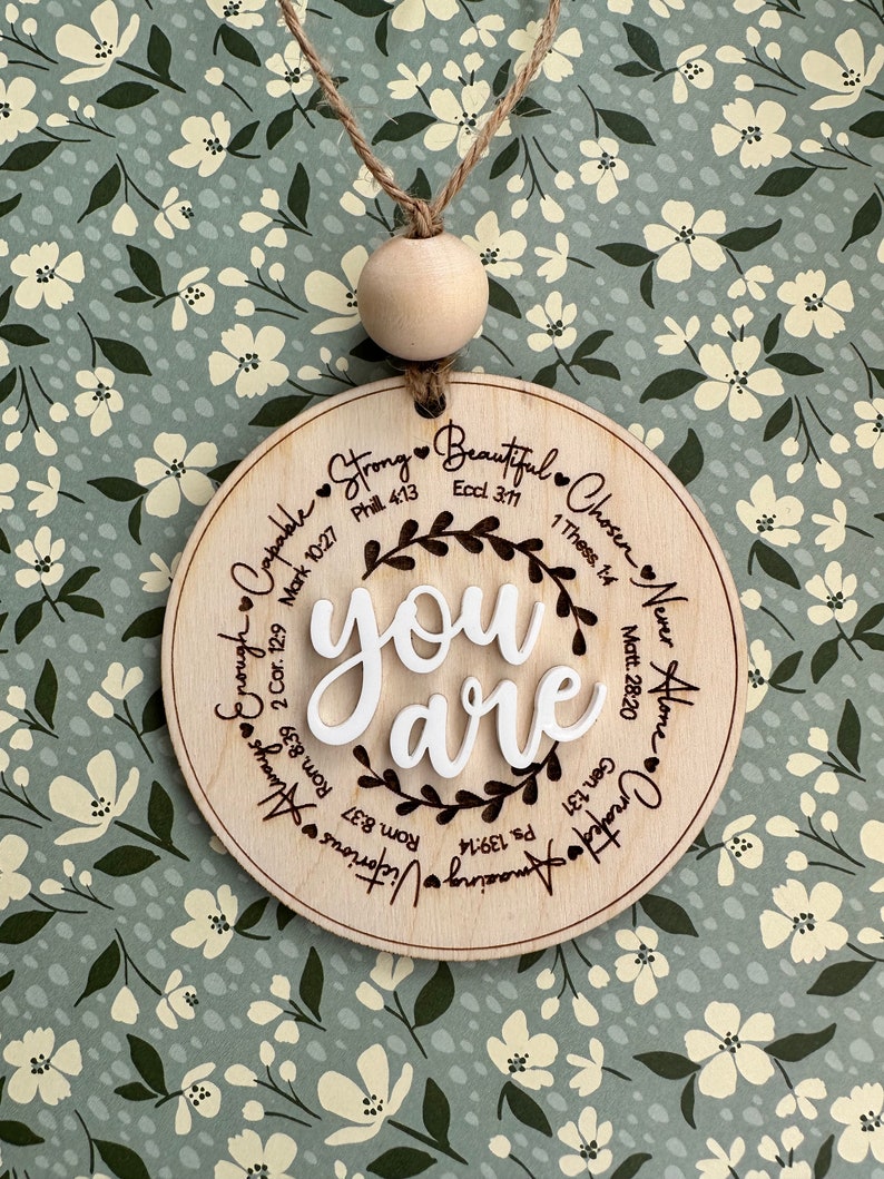 May include: A round wooden ornament with the words "you are" in white, surrounded by inspirational words and Bible verses. The ornament hangs from a twine string with a wooden bead.