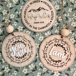 May include: Three wooden circular ornaments with inspirational quotes. One reads "Waymaker" with a mountain graphic, another says "You Are", and the last says "I Am". Each has a jute string and wooden bead for hanging.