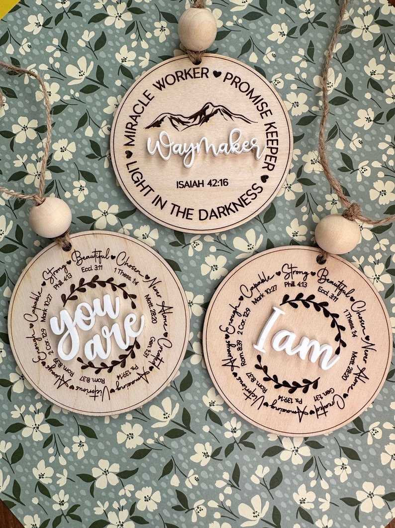 May include: Three wooden circular ornaments with inspirational quotes. One reads "Waymaker" with a mountain graphic, another says "You Are", and the last says "I Am". Each has a jute string and wooden bead for hanging.