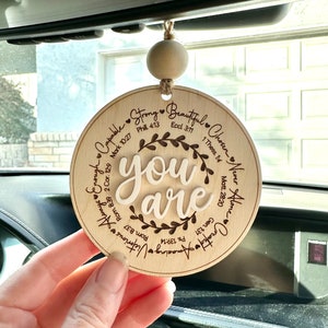 May include: A round, wooden car charm with the words "you are" in a cursive font, surrounded by inspirational words and Bible verses. The charm is suspended by a natural-colored wooden bead and twine.