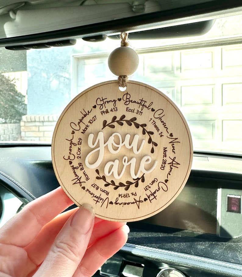 May include: A round, wooden car charm with the words "you are" in a cursive font, surrounded by inspirational words and Bible verses. The charm is suspended by a natural-colored wooden bead and twine.