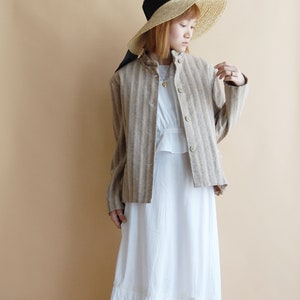 Op de afbeelding: Een vrouw draagt een gestreept bruin en wit jasje met een witte jurk en een brede strohoed met een zwart lint.