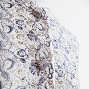 May include: A close-up of a cream-colored fabric with a delicate floral embroidery pattern in shades of brown and gray. Two silver buttons with a light purple center are visible on the fabric.