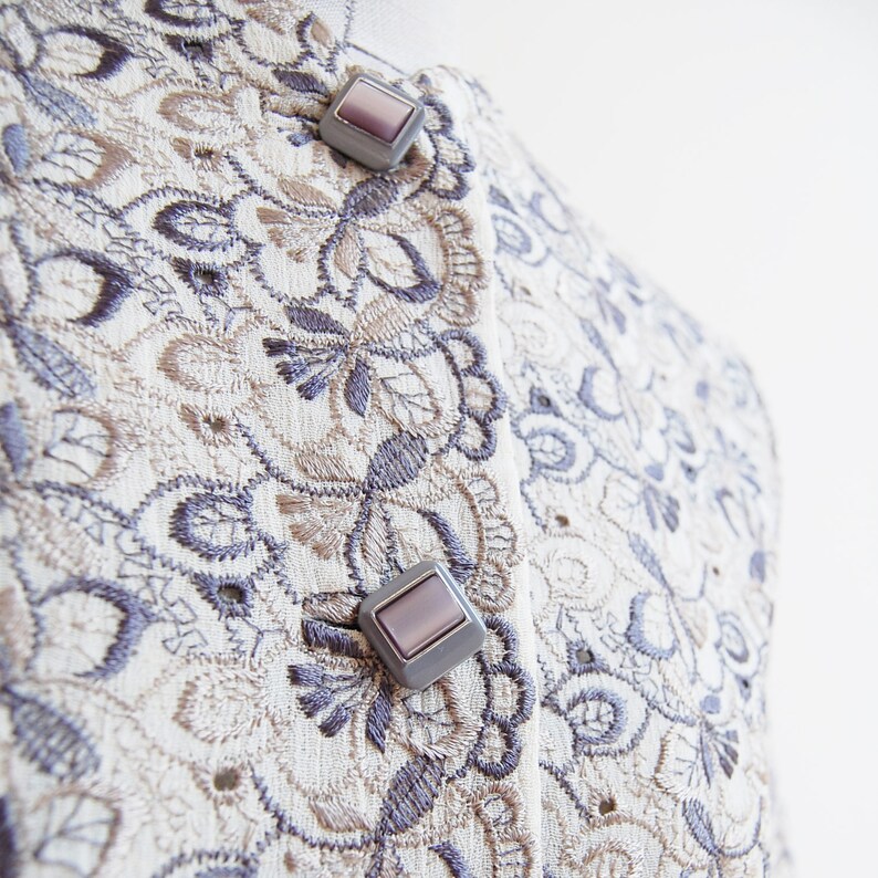 May include: A close-up of a cream-colored fabric with a delicate floral embroidery pattern in shades of brown and gray. Two silver buttons with a light purple center are visible on the fabric.