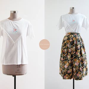以下が含まれることがあります： 白い半袖Tシャツに花柄の刺繍が施されています。Tシャツは黒地に花柄のプリントが施されたスカートと組み合わされています。