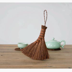 May include: A brown and natural fiber broom with a long handle and orange accents. The broom is sitting on a wooden table with a green tea pot and a small bowl.