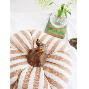 May include: A white and brown striped fabric pumpkin with a brown wooden stem. The pumpkin is sitting on a white surface.