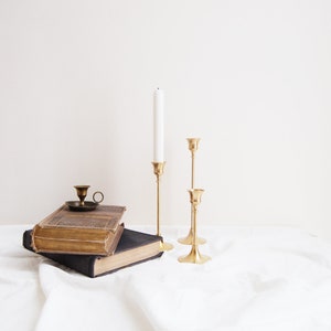 May include: Three gold candle holders with a white candle in the tallest holder. The candle holders are on a white surface with a stack of two books.