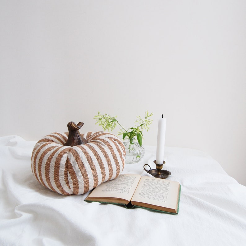May include: A striped brown and white pumpkin-shaped pillow sits on a white linen tablecloth with a book and a candle holder.