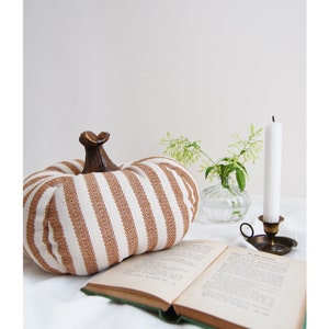 May include: A brown and white striped fabric pumpkin with a wooden stem sits on a white surface next to a book and a candle holder with a white candle.