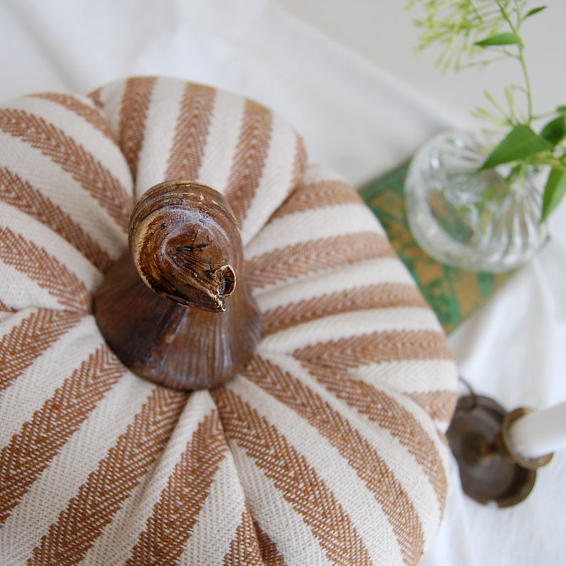 May include: A white and brown striped fabric pumpkin with a brown wooden stem. The pumpkin is sitting on a white surface.